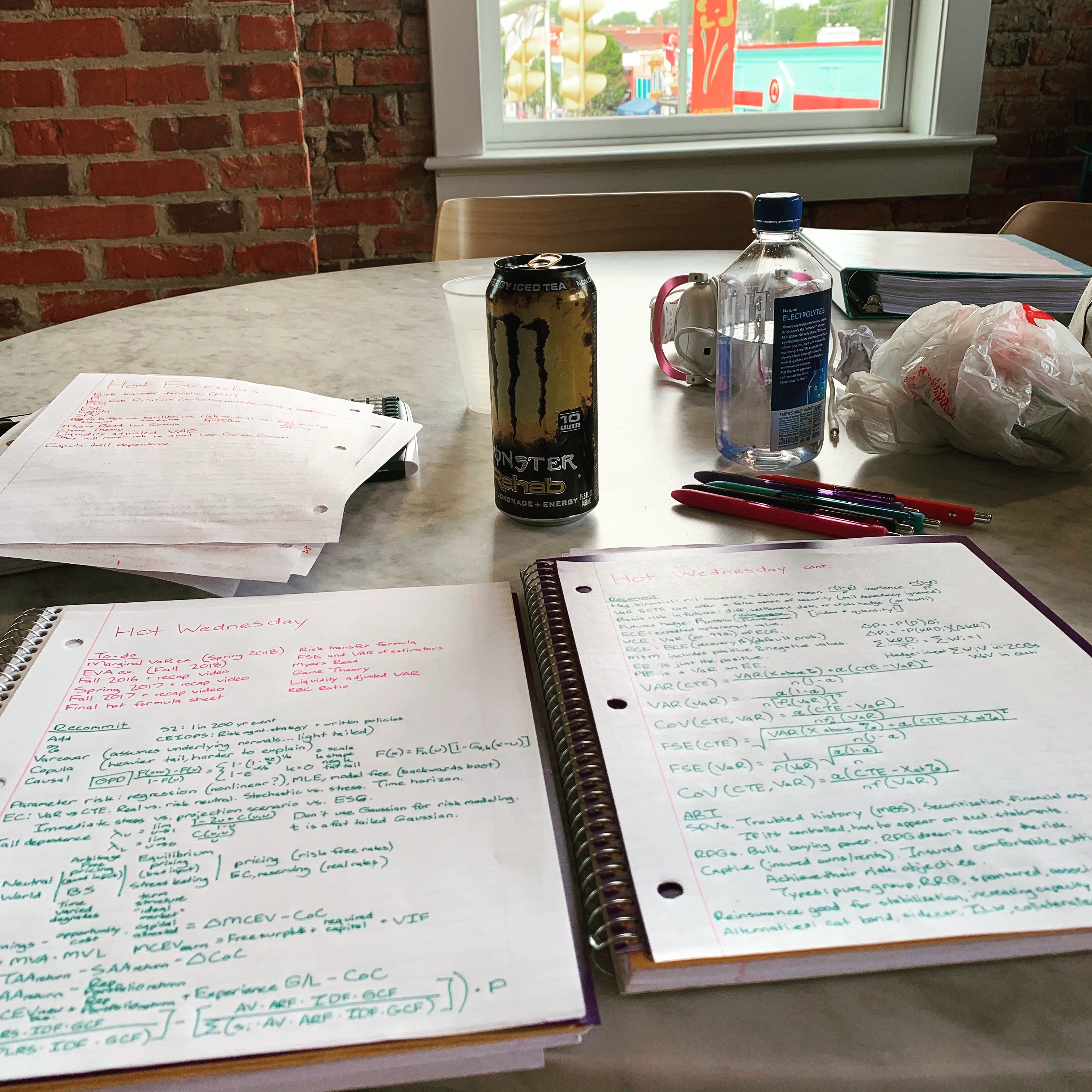 A naturally lit, white marble table at the Capital One Café in Richmond, VA. The table is covered in study notes and materials, and other important study tools like colored gel pens and headphones. The energy drink indicates crunch time — exam day is approaching.